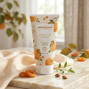A tube of Herbsalot Apricot Scrub is displayed on a marble surface with a soft cream fabric draped beside it. The packaging features apricot illustrations and text highlighting gentle exfoliation for smooth, radiant skin. Dried apricots, almonds, and green leaves are arranged around the product, creating a fresh, natural, and elegant skincare presentation.